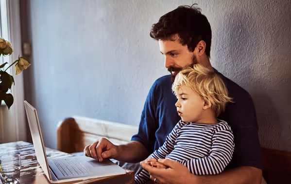 Een man zit met zijn zoon op schoot terwijl ze samen naar zijn laptopscherm kijken.